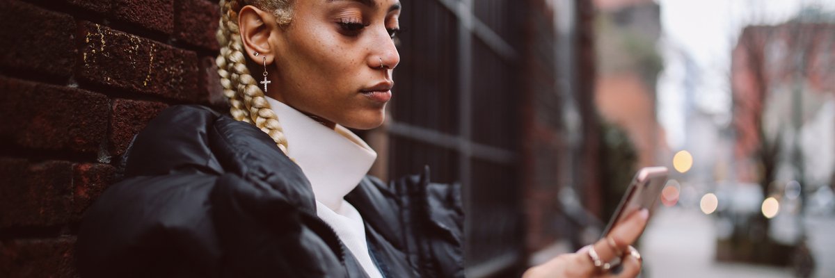 A young adult uses their phone while leaning against a brick wall in a city.