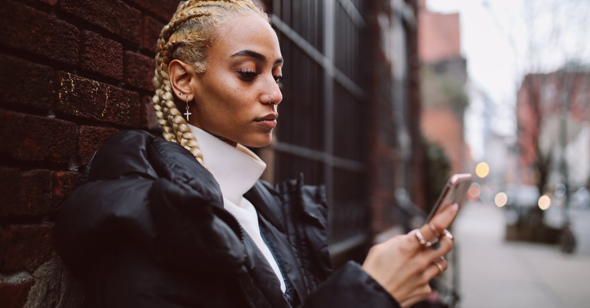 A young adult uses their phone while leaning against a brick wall in a city.