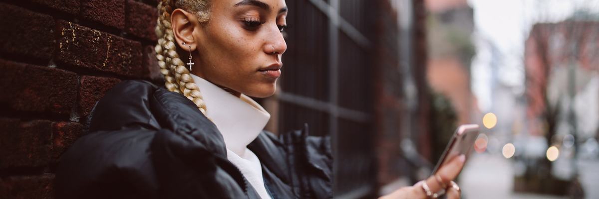 A young adult uses their phone while leaning against a brick wall in a city.