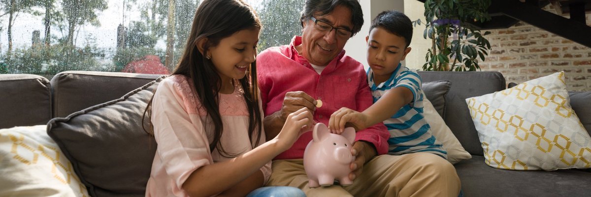 Dad sitting on couch with his two young kids as they all put coins into a piggy bank.