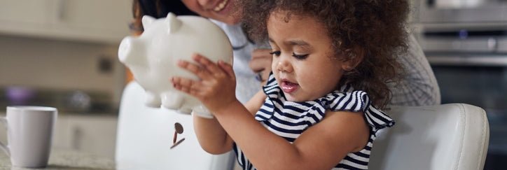 Child shakes money out of a piggy bank as the parent watches and smiles.