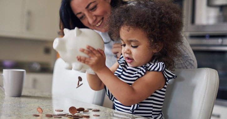 Child shakes money out of a piggy bank as the parent watches and smiles.