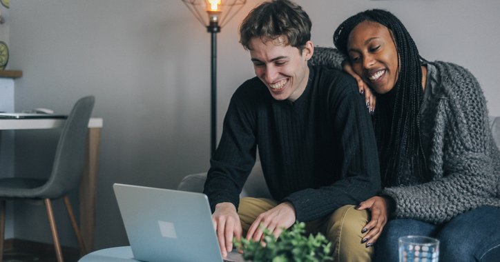 A smiling couple sitting on the couch while looking something up on their laptop.
