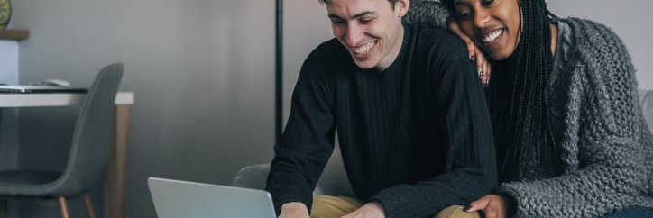 A smiling couple sitting on the couch while looking something up on their laptop.