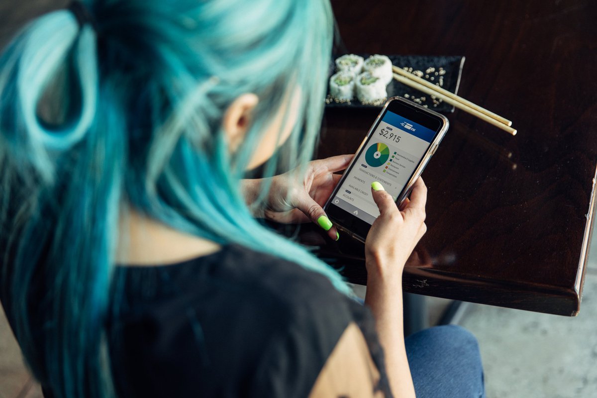 Over-the-shoulder view of a blue-haired young woman checking her finances on her phone.