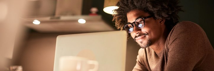 A man standing in his kitchen lit by the glow of his laptop screen.