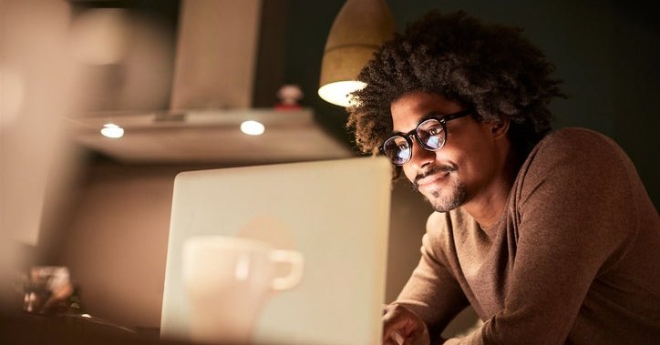 A man standing in his kitchen lit by the glow of his laptop screen.
