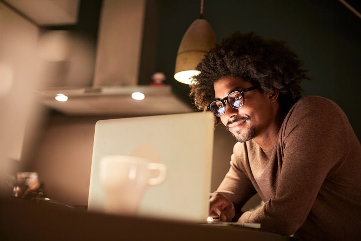 A man standing in his kitchen lit by the glow of his laptop screen.