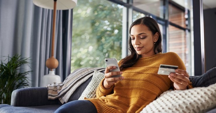Woman with credit card in her hand using cellphone while reclining on sofa.