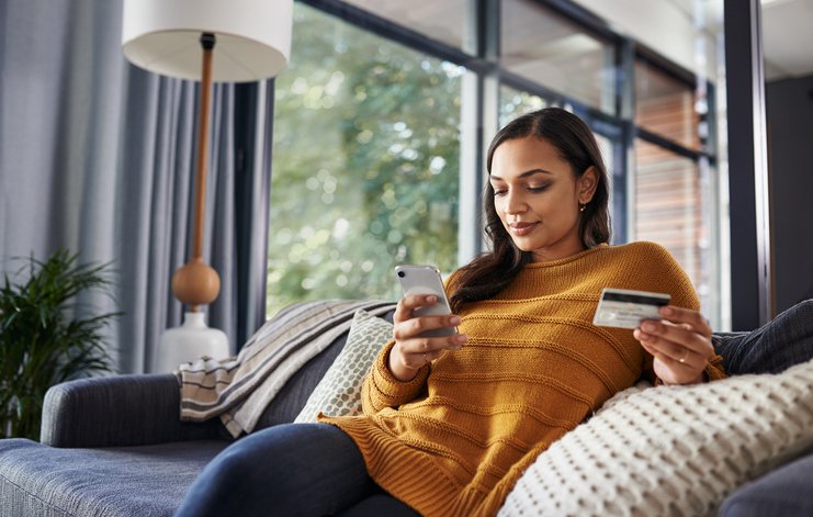 Woman with credit card in her hand using cellphone while reclining on sofa.