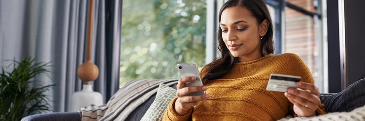 Woman with credit card in her hand using cellphone while reclining on sofa.