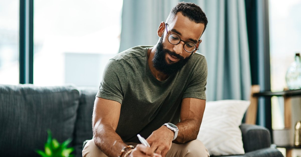 A buff, bearded, bespectacled young man looking relaxed and confident while marking something down on a piece of paper.