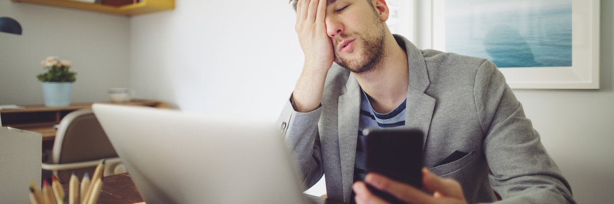 Young man puts his hand to his face in frustration while holding his phone and looking at laptop.