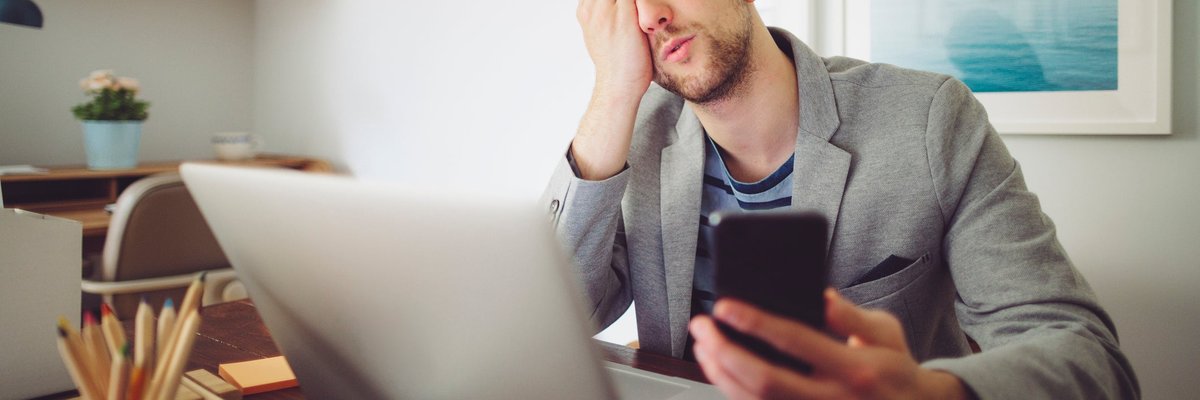 Young man puts his hand to his face in frustration while holding his phone and looking at laptop.