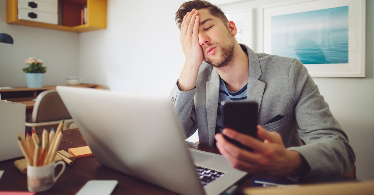 Young man puts his hand to his face in frustration while holding his phone and looking at laptop.