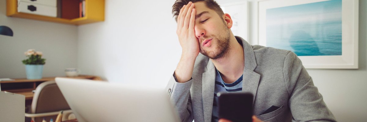 Young man puts his hand to his face in frustration while holding his phone and looking at laptop.