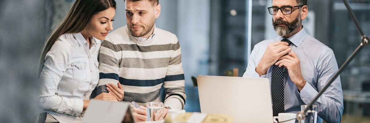 A young couple looks unsure about signing a financial document in a financer's office.