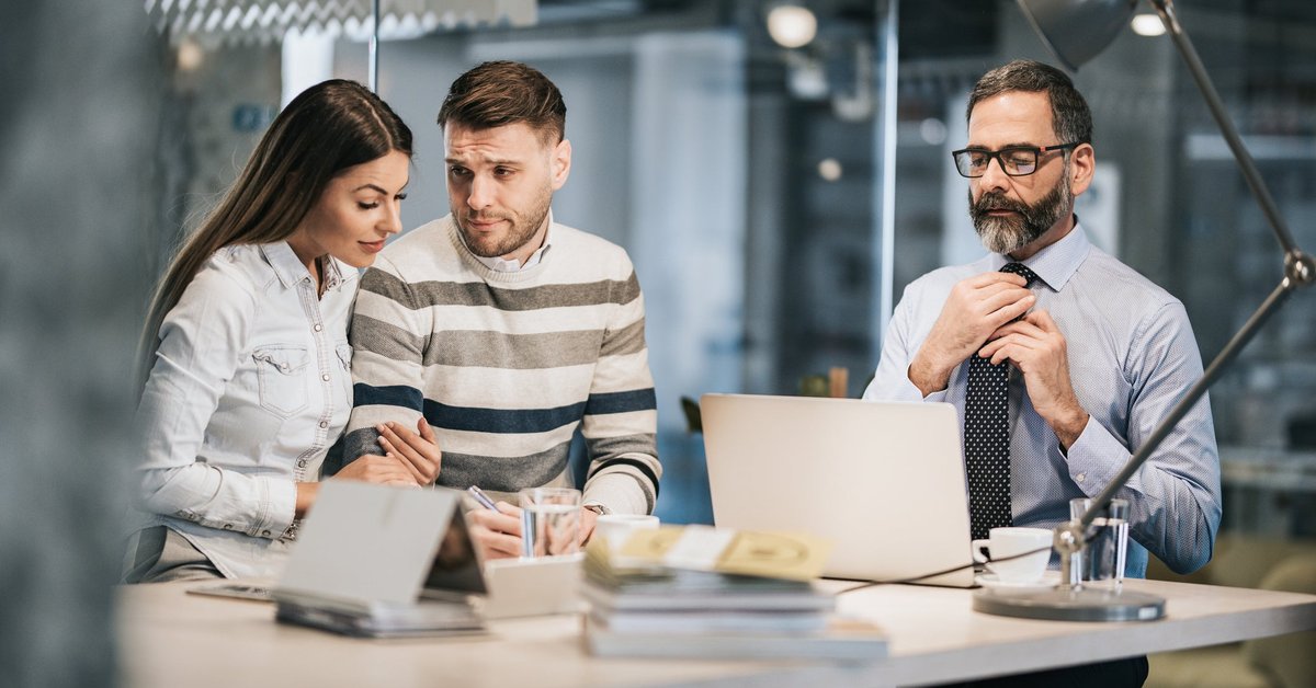 A young couple looks unsure about signing a financial document in a financer's office.