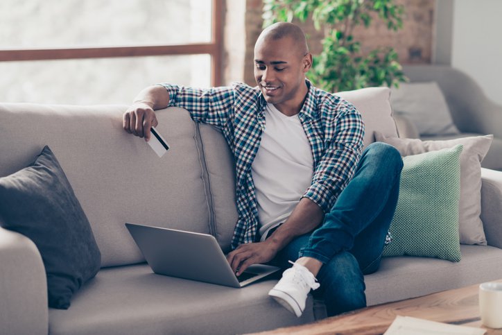 Smiling man sitting on sofa with laptop and credit card.