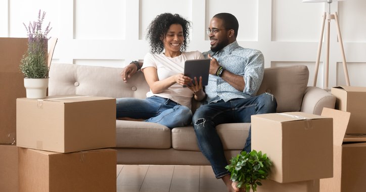 Couple sits on sofa looking at tablet and surrounded by boxes.