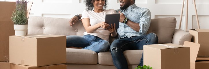 Couple sits on sofa looking at tablet and surrounded by boxes.
