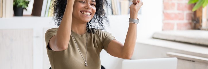 Smiling woman sitting at computer raises both hands in celebration.