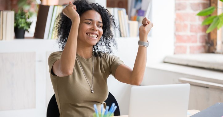 Smiling woman sitting at computer raises both hands in celebration.
