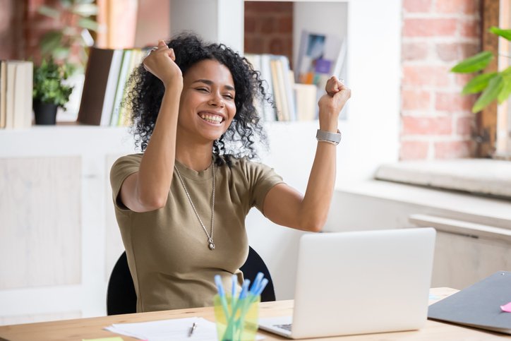 Smiling woman sitting at computer raises both hands in celebration.