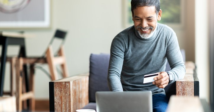 A man sitting in an armchair and typing into his laptop while holding a credit card in one hand.
