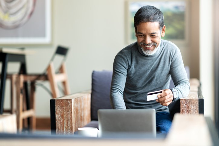 A man sitting in an armchair and typing into his laptop while holding a credit card in one hand.