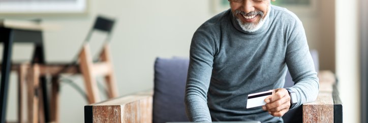 A man sitting in an armchair and typing into his laptop while holding a credit card in one hand.