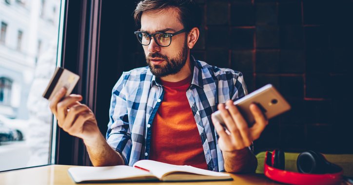 Man in blue checked shirt looks thoughtful with phone in one hand and credit card in the other.
