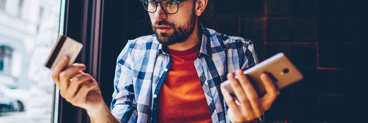 Man in blue checked shirt looks thoughtful with phone in one hand and credit card in the other.