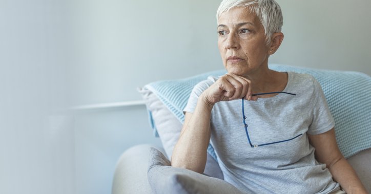 Grey-haired woman holding glasses rests hand on chin and looks thoughtful.