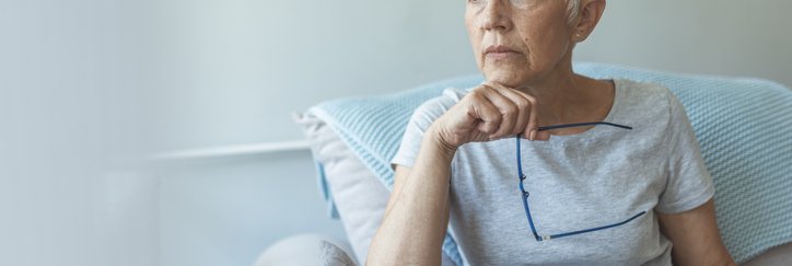 Grey-haired woman holding glasses rests hand on chin and looks thoughtful.