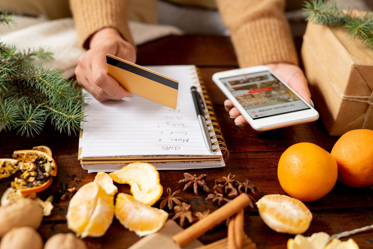 A pair of hands holds a credit card and phone over a notebook and some holiday garlands and chestnuts and fruits.