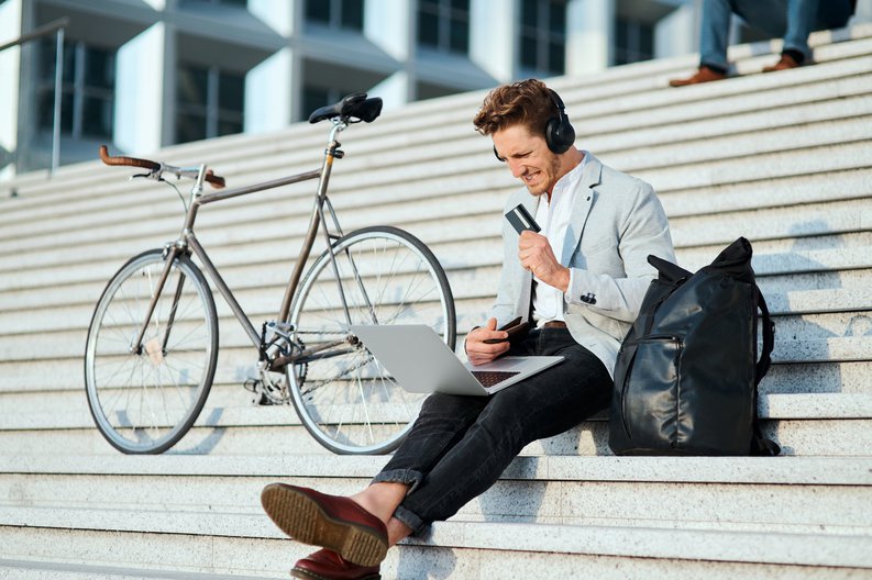 Young man sitting on staircase holding credit while looking at laptop and grimacing.