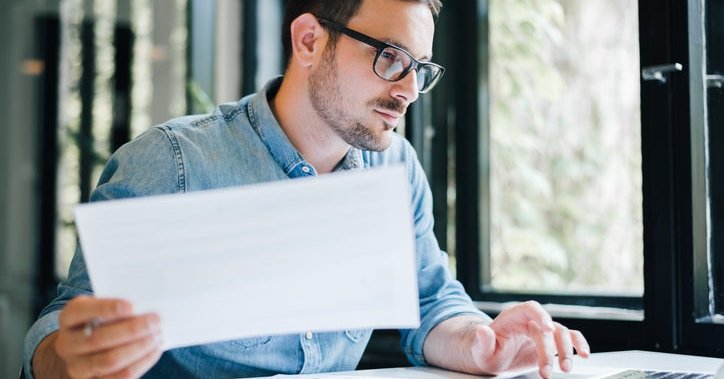Man at desk holding document in one hand and typing with the other.