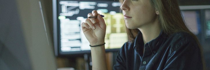 A young woman sits at a desk surrounded by monitors displaying financial data.