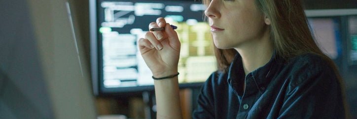 A young woman sits at a desk surrounded by monitors displaying financial data.