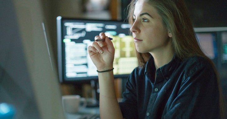 A young woman sits at a desk surrounded by monitors displaying financial data.