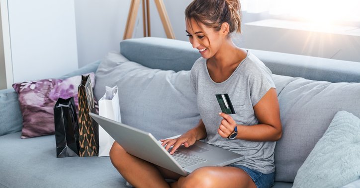 Smiling woman with laptop sits cross legged on sofa holding a credit card.