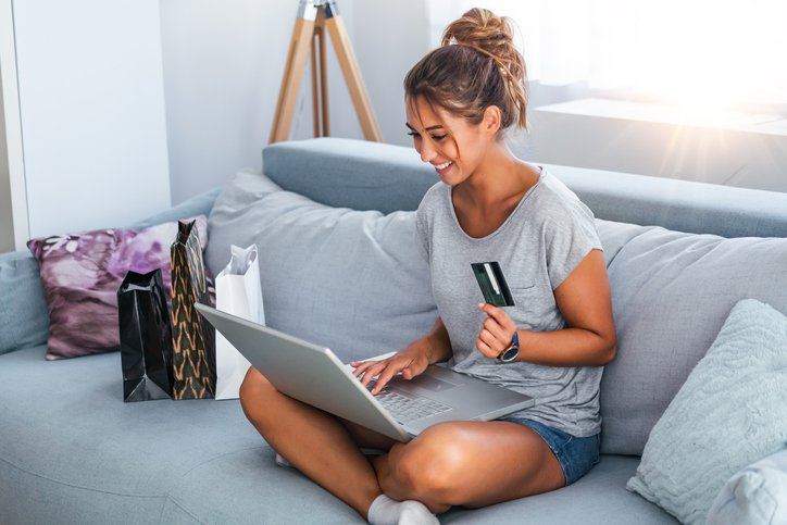 Smiling woman with laptop sits cross legged on sofa holding a credit card.