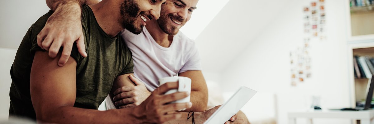 A couple sits in bed drinking coffee and smiling at something on a laptop screen.