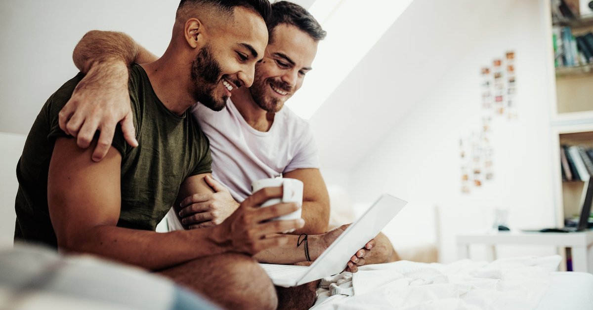 A couple sits in bed drinking coffee and smiling at something on a laptop screen.