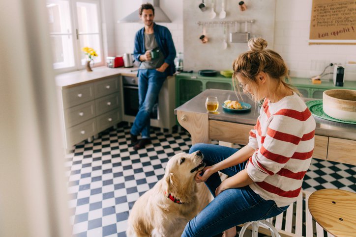 Woman with dog in kitchen foreground, while man looks on.