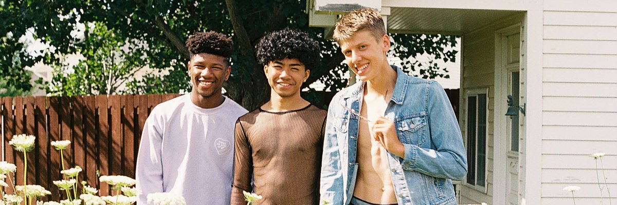 Three young adults stand together in a field of flowers in front of a modest house.