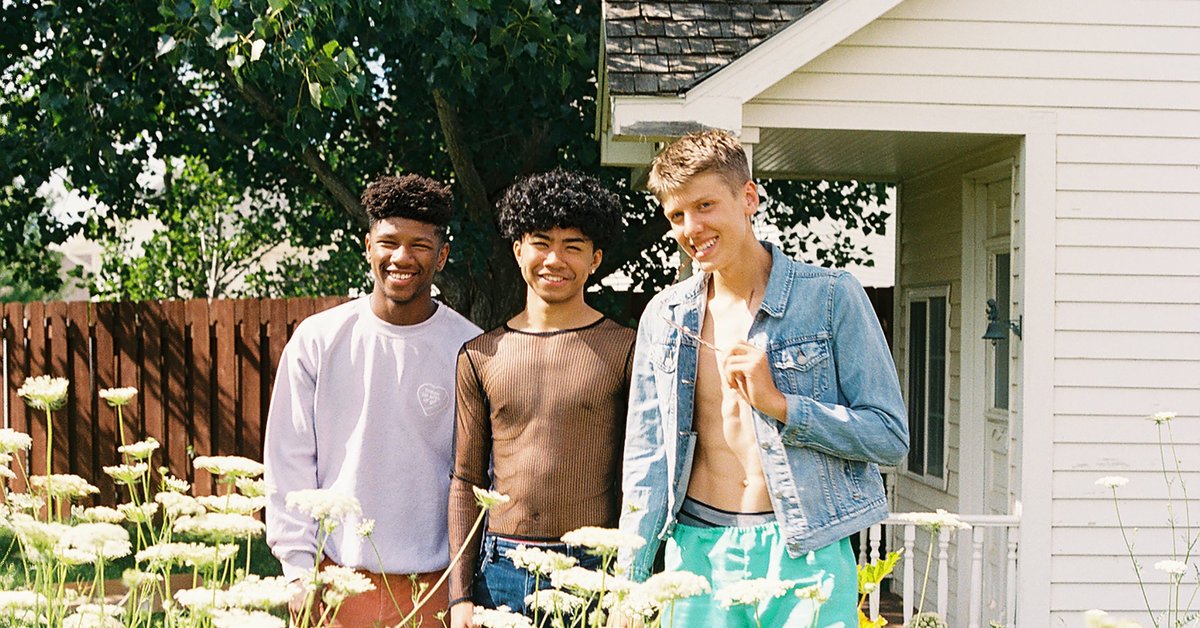 Three young adults stand together in a field of flowers in front of a modest house.