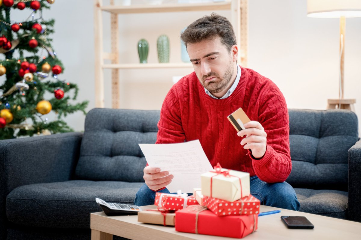 Man in Christmas sweater sitting in front of a pile of gifts while holding a credit card and looking at a document.