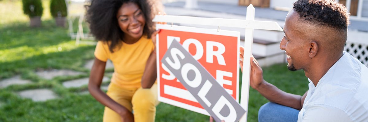 A young couple happily attaches a "Sold" banner to a "For Sale" sign outside a house.